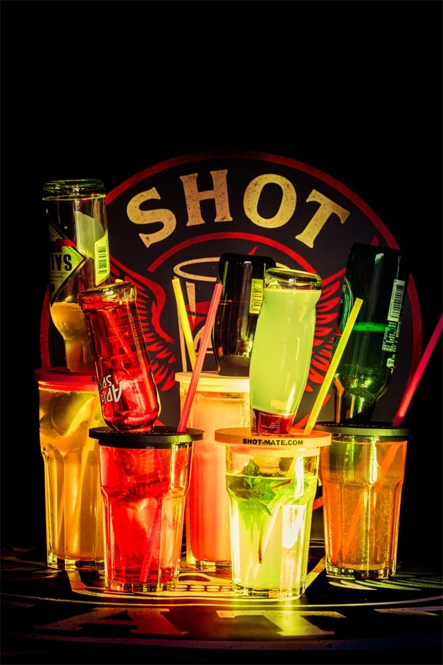 Fünf bunte Gläser mit eingesetzten Flaschen und Shot-Mate Booze Dock Aufsätzen vor beleuchtetem Logo.