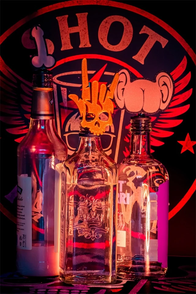 Drei Flaschen mit Shot-Mate-Aufsätzen in Penis-, Rockerhand- und Po-Form vor beleuchtetem Markenlogo.