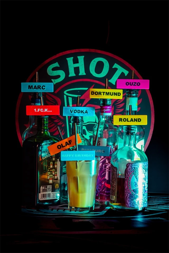 Flaschen und Glas mit bunten Shot-Mate-Clips in Neonfarben vor großem Markenlogo.